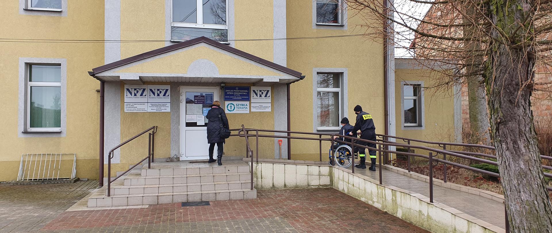 Na tle budynku przychodni lekarskiej obecne są trzy osoby. Z prawej strony widoczni na podjeździe dla osób niepełnosprawnych są dwaj mężczyźni: jeden będący na wózku inwalidzkim pchanym przez strażaka. Drzwi budynku otwiera im kobieta. 