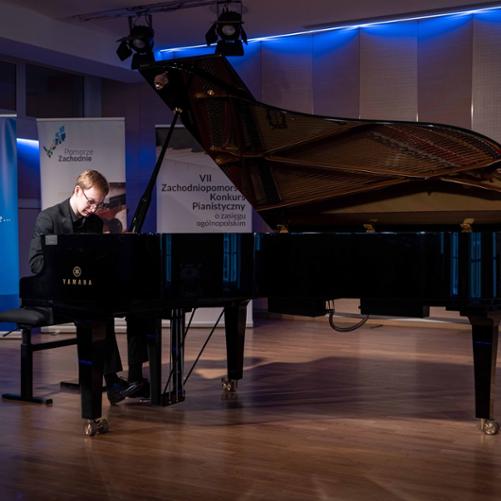 Zdjęcie przedstawia wykonawcę grającego na fortepianie na scenie sali koncertowej, w tle banery Radia Szczecin, Pomorza Zachodniego oraz konkursu