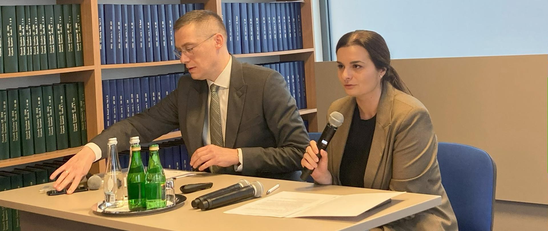 W sali konferencyjnej przy stole siedzi mężczyzna oraz kobieta, która mówi do trzymanego w ręku mikrofonu