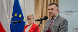 Zdjęcie z boku, na sali stoi minister Wieczorek i wiceminister Mrówczyńska w czerwonym garniturze, minister trzyma w ręku czerwoną teczkę i mówi do mikrofonu na stojaku.