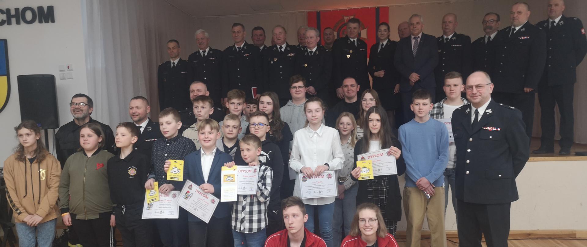 W sali gminnego ośrodka kultury stoją zwycięzcy eliminacji powiatowych OTWP. Dzieci trzymają w rękach nagrody oraz dyplomy. Za nimi stoją przedstawiciele władz oraz Komendant Powiatowy PSP w Bytowie