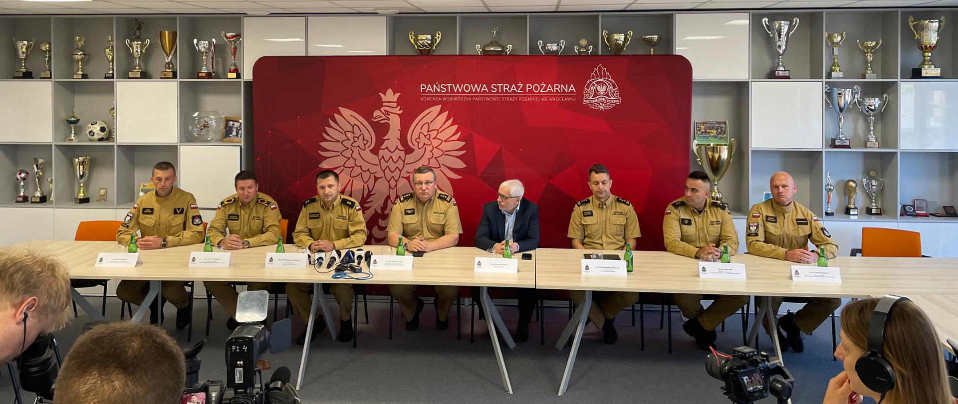 Strażacy wraz z wojewodą dolnośląskim siedzący przy stole.