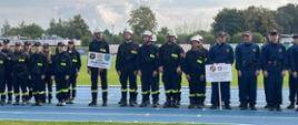 Strażacy ochotnicy w czarnych ubraniach koszarowych stojący w jednej linii i czekający na rozpoczęcie zawodów
