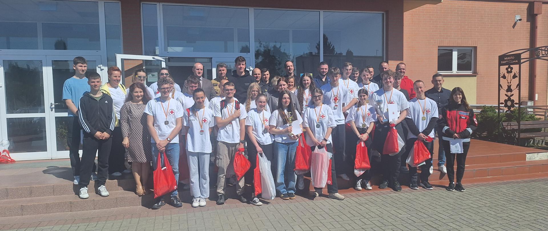 Grupa osób stoi przed budynkiem. Osoby z przodu mają na sobie białe koszulki. W grupie znajdują się również strażacy ubrani w czarne umundurowanie, Starosta Tucholski oraz Komendant Powiatowy PSP w Tucholi. Osoby z przodu trzymają czerwone i białe torby. W tyle widać okna budynku. Budynek jest koloru pomarańczowego. Część osób stoi na schodach przed budynkiem. 