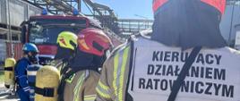Strażacy w mundurach i hełmach. Jeden ubrany w kamizelkę z napisem "KIERUJĄCY DZIAŁANIEM RATOWNICZYM".
