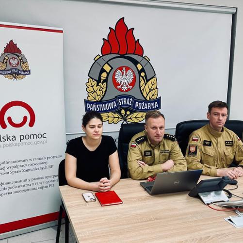 3 mężczyzn w mundurach i kobieta siedzą przygotowując się do zajęć online. Z tyłu baner z logo Polskiej pomocy i Państwowej Straży Pożarnej