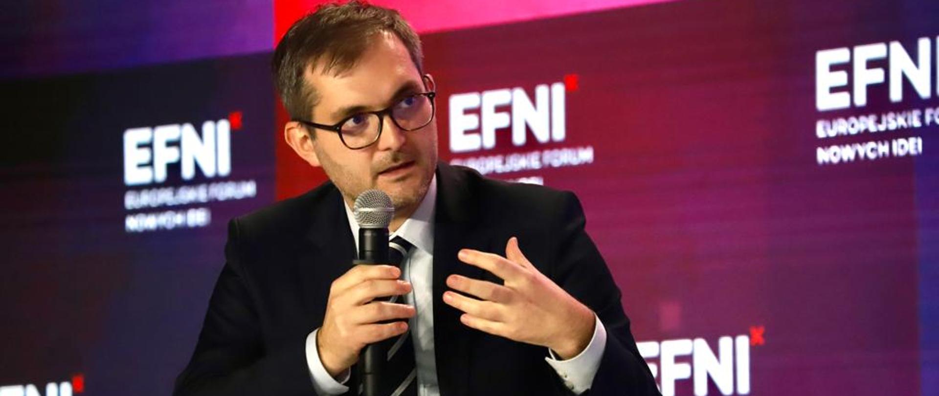 Wiceminister rozwoju i technologii Marek Niedużak trzymający mikrofon w dłoni podczas obrad EFNI'21 w Sopocie, za jego plecami baner w kolorach czerwieni, niebieskiego i granatu z logo EFNI