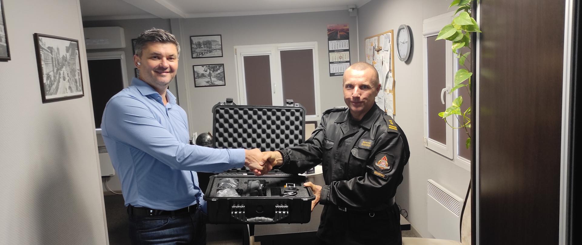 Na zdjęciu z lewej strony w niebieskiej koszuli Członek Zarządu Spółki Pan Aleksander Iwanowicz oraz z prawej bryg. Ireneusz Ścibiorek. Ściskają dłonie pokazując sprzęt w czarnej walizce.