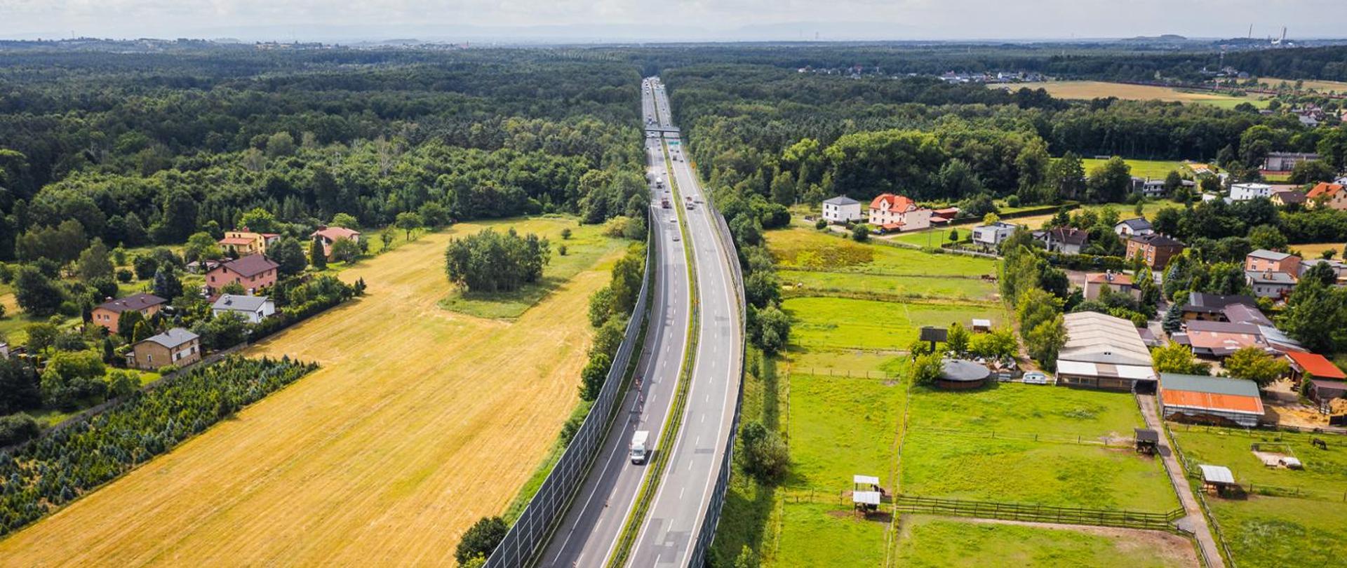 Widok z lotu ptaka na drogę ekspresową S1 przebiegającą między terenami rolnymi, zabudową jednorodzinną i lasami. Wzdłuż trasy znajdują się wysokie ekrany akustyczne, a po jezdni poruszają się samochody ciężarowe i osobowe.