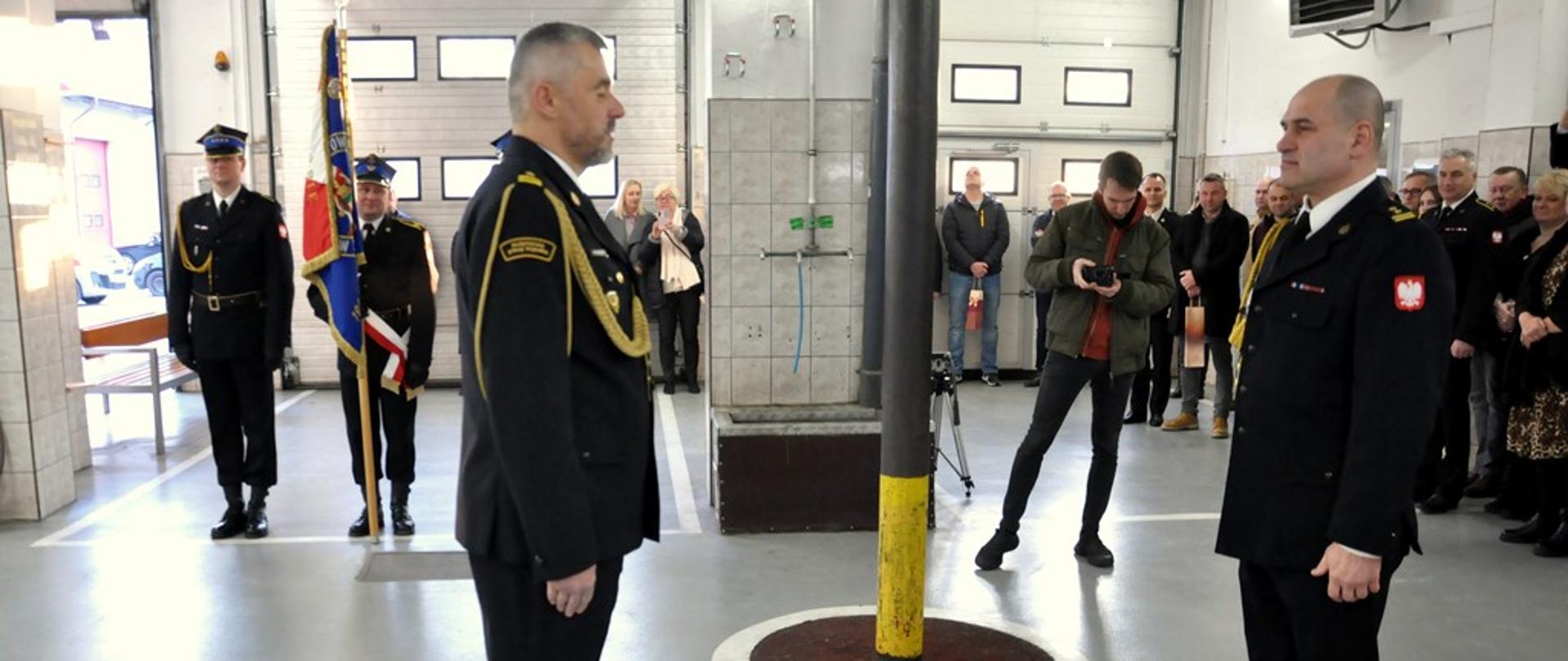 Pomieszczenie garażowe. Na pierwszym planie stoi naprzeciw siebie, w pozycji zasadniczej dwóch funkcjonariuszy PSP w umundurowaniu wyjściowym, bez rogatywki. Z lewej strony stoi poczet sztandarowy, z lewej strony stoi pod ścianą grupa osób cywilnych.