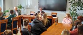 Grupa uczniów siedzących przy stołach i słuchających wypowiedzi jednego z dwóch starszych mężczyzn.
