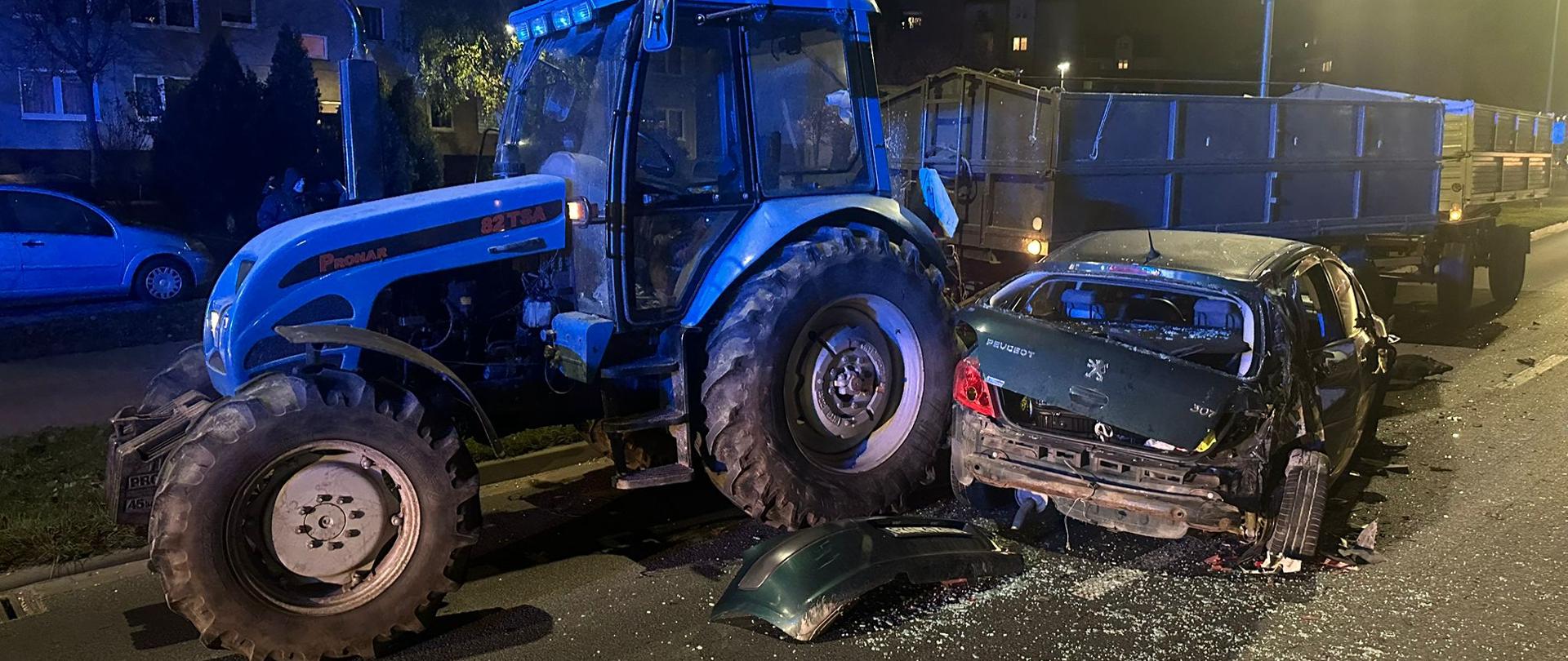 Na zdjęciu widać miejsce wypadku drogowego nocą. Na oświetlonej ulicy stoi niebieski traktor z przyczepą, który uderzył w tył ciemnozielonego samochodu osobowego marki Peugeot. Auto jest poważnie zniszczone z tyłu, wokół leżą fragmenty karoserii i szkło. W tle widać blok mieszkalny z zapalonymi światłami w oknach oraz zaparkowane samochody.