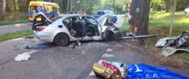 Wrak samochodu osobowego marki BMW na ulicy w odległości 1 metr od drzewa w które uderzyło. Na pierwszym planie torba medyczna na macie sprzętowej koloru niebieskiego. W tle karetka pogotowia ratunkowego, radiowóz wraz z funkcjonariuszami Policji.