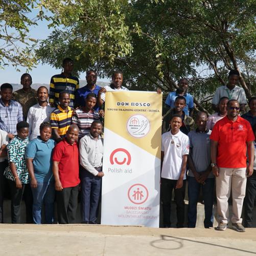 Pod drzewem stoją w trzech rzędach nauczyciele i nauczycielki, z których część trzyma baner informacyjny szkoły