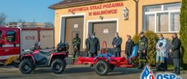 Kolorowa fotografia wykonana w słonecznym dniu przedstawia na pierwszym planie nowy czerwono-czarny pojazd quad z przyczepką na placu przed remizą Ochotniczej Straży Pożarnej. W tle od lewej: czerwony lekki pojazd ratowniczo-gaśniczy z widocznym czerwonym numerem operacyjnym 417R36 na zabudowie skrytki pojazdu. Pozostałe tło wypełnia elewacja frontowa budynku z widocznymi dwoma brązowymi bramami wyjazdowymi, dużymi białymi oknami i czerwonym napisem na elewacji o treści: Ochotnicza Straż Pożarna w Malinówce. Na elewacji widoczna figura w gablotce. Przed budynkiem stoją osoby cywilne, umundurowani strażacy i osoba duchowna w komży. Na dole fotografii naniesione niebiesko-czerwone logo z napisem OSP.pl.