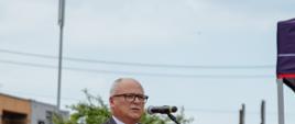 Starosta w garniturze przemawia do mikrofonu przy mównicy z logo Państwowej Straży Pożarnej podczas oficjalnej uroczystości plenerowej.