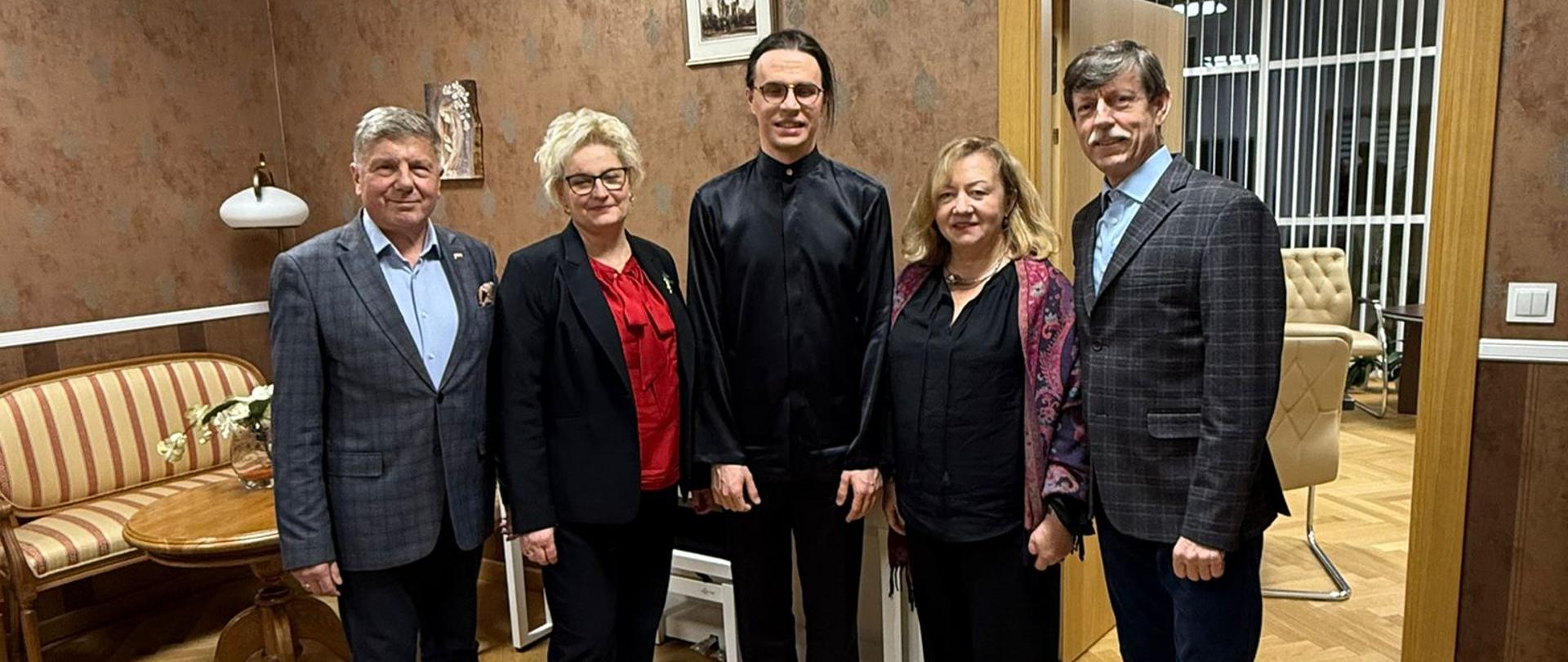 Na zdjęciu widoczna jest grupa pięciu dorosłych osób stojących obok siebie w gabinecie lub sali spotkań. Wszyscy są elegancko ubrani – kobiety w strojach wizytowych, mężczyźni w marynarkach; jedna z osób ma na sobie czarny strój koncertowy. Wnętrze ma klasyczny charakter: drewniana podłoga, tapicerowane fotele, stolik kawowy, regał z dokumentami oraz obrazy na ścianach. W tle widać drzwi prowadzące do sąsiedniego pomieszczenia. Osoby pozują do wspólnego pamiątkowego zdjęcia po wydarzeniu kulturalnym.