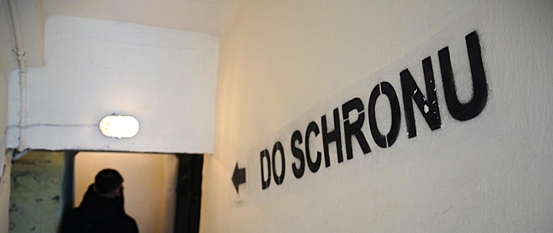 Ściana ze strzałką i napisem do schronu. W tle człowiek wchodzący do schronu 