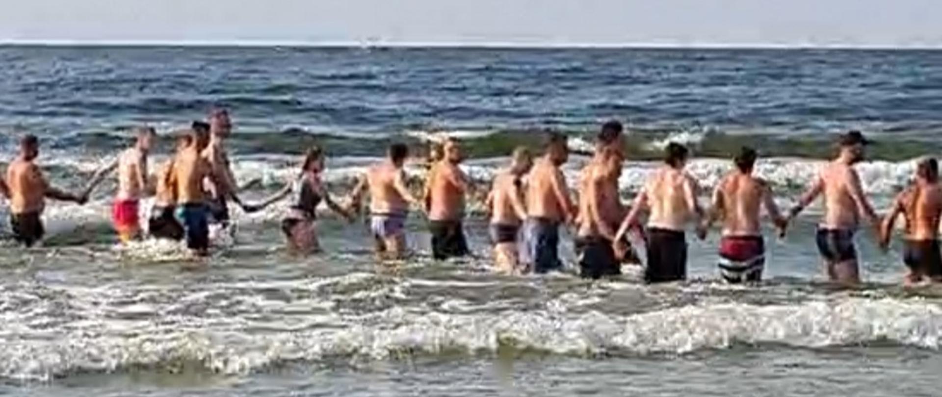 Na zdjęciu widać ludzi w wodzie trzymających się za ręce.