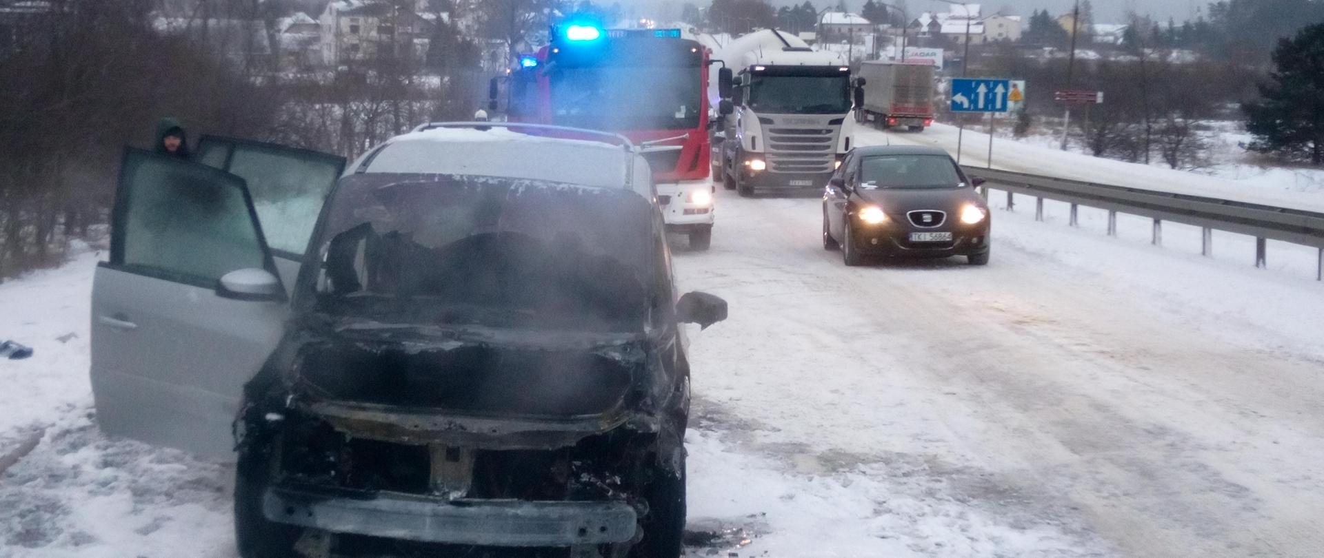 Na zdjęciu znajduje się spalony samochód marki Renault. Ma otwarte prawe drzwi - z jego wnętrza wydobywa się jeszcze dym. W tle
samochód straży pożarnej na sygnałach. Jezdnia jest cała w śniegu.
