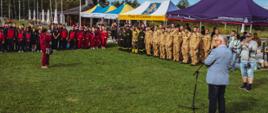 Fotografia zrobiona z góry z szerokiego kąta przedstawia ceremonię strażacką, podczas której starosta wodzisławski stoi plecami do widza. Przemawia on przez stojący przed nim mikrofon do uformowanej w trzech szeregach, ubranej w mundury grupy strażaków. Inna uformowana grupa ubranej w dresy młodzieży stoi z boku. Za strażakami widać rozstawione namioty, na których napisane jest „Powiat Wodzisławski” oraz „Państwowa Straż Pożarna”. Tło ujęte na zdjęciu to otoczony drzewami las, z którego wyłania się letni budynek. Zdjęcie ma wyraźnie podniesioną jasność, co dodaje mu dynamicznego charakteru.