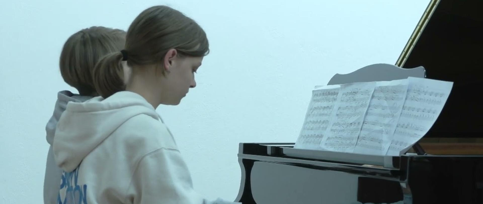 Zdjęcie przedstawia dwoje uczniów grających na czarnym fortepianie na sali koncertowej w Państwowej Szkole Muzycznej I stopnia w Bełchatowie.