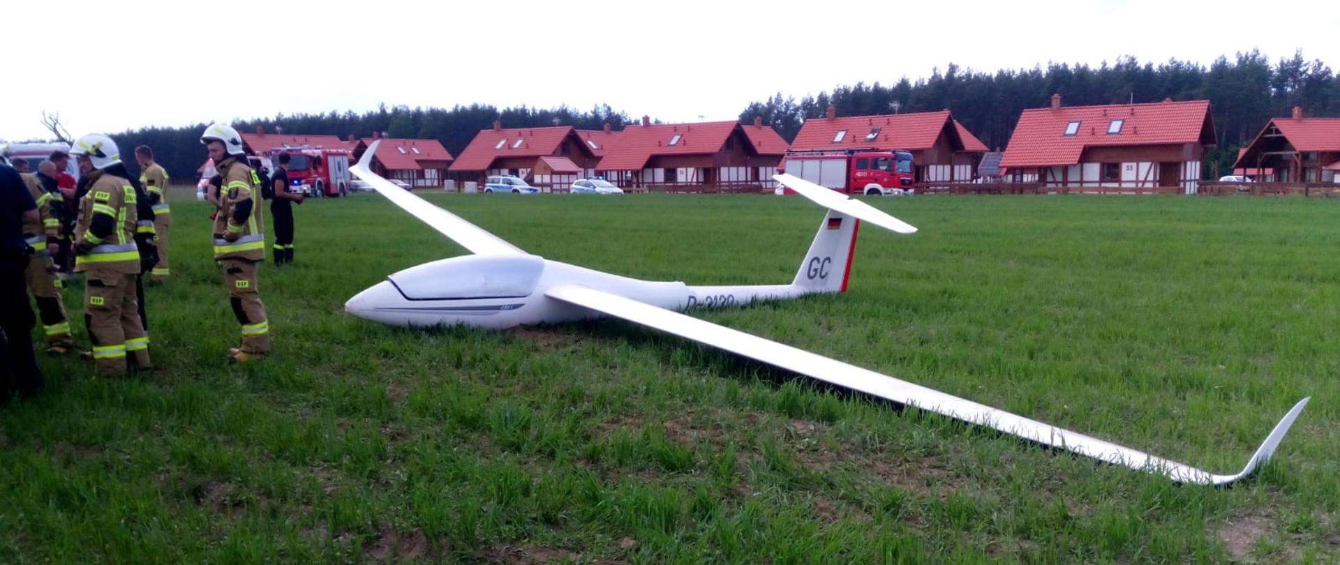 Na zdjęciu znajduje się biały jednoosobowy szybowiec na polu uprawnym. Przed szybowcem stoją umundurowani strażacy.