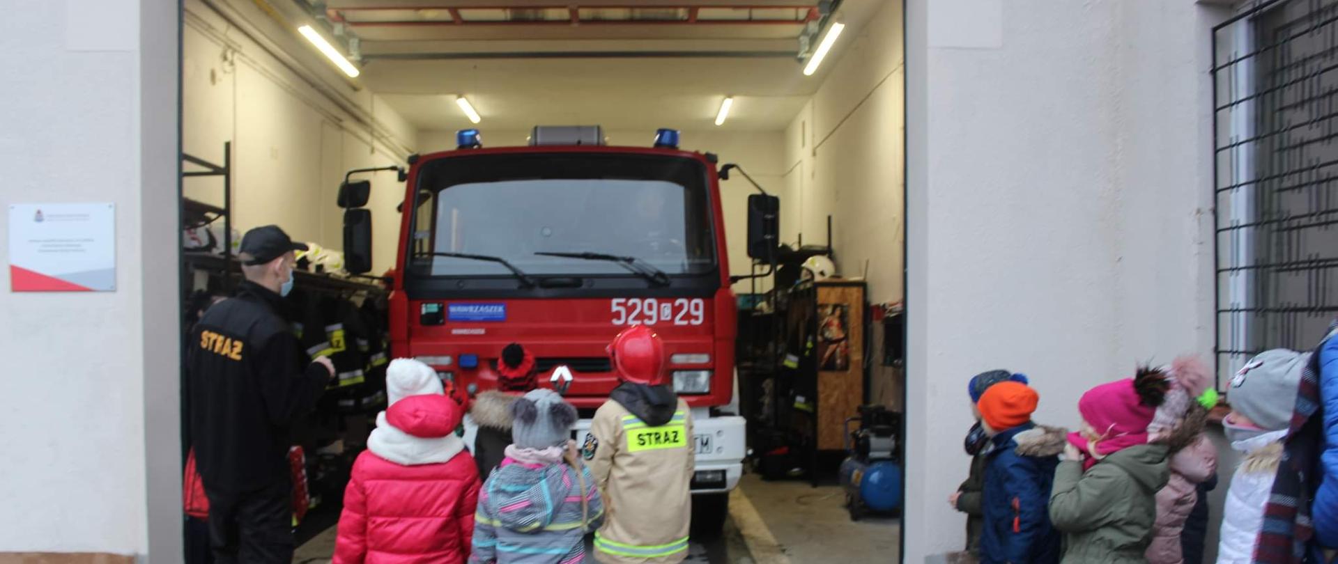 Zdjęcie wykonano za dnia przed remizą Ochotniczej Straży Pożarnej w Kamieniu Krajeńskim. Otwarta brama wjazdowa garażu, w garażu samochód gaśniczy marki Renault, przed bramą stoi strażak Państwowej Straży Pożarnej ubrany w mundur koszarowy, po jego prawej stronie grupa dzieci w kurtkach i czapkach zimowych.