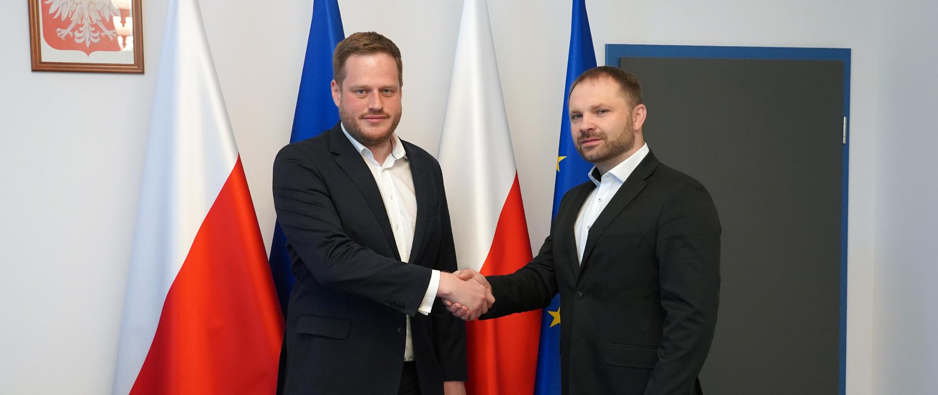 Minister Janusz Cieszyński i nowy dyrektor Centralnego Ośrodka Informatyki Przemysław Koch na tle flag Polski i Unii Europejskiej.