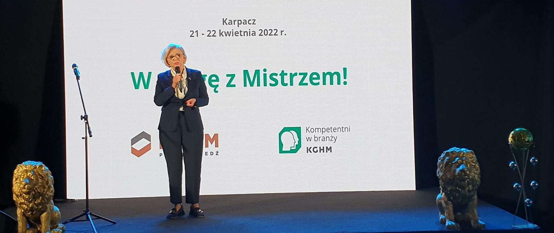 Na scenie do mikrofonu przemawia wiceminister Marzena Machałek. Ubrana w granatowy garnitur i żółtą bluzkę. Po obydwu stronach sceny widać posągi przedstawiające lwy. W tle napis Karpacz oraz data 21-22 kwietnia oraz zielony napis pod datą W drogę z mistrzem W drogę z mistrzem – wydarzenie z udziałem minister Marzeny Machałek