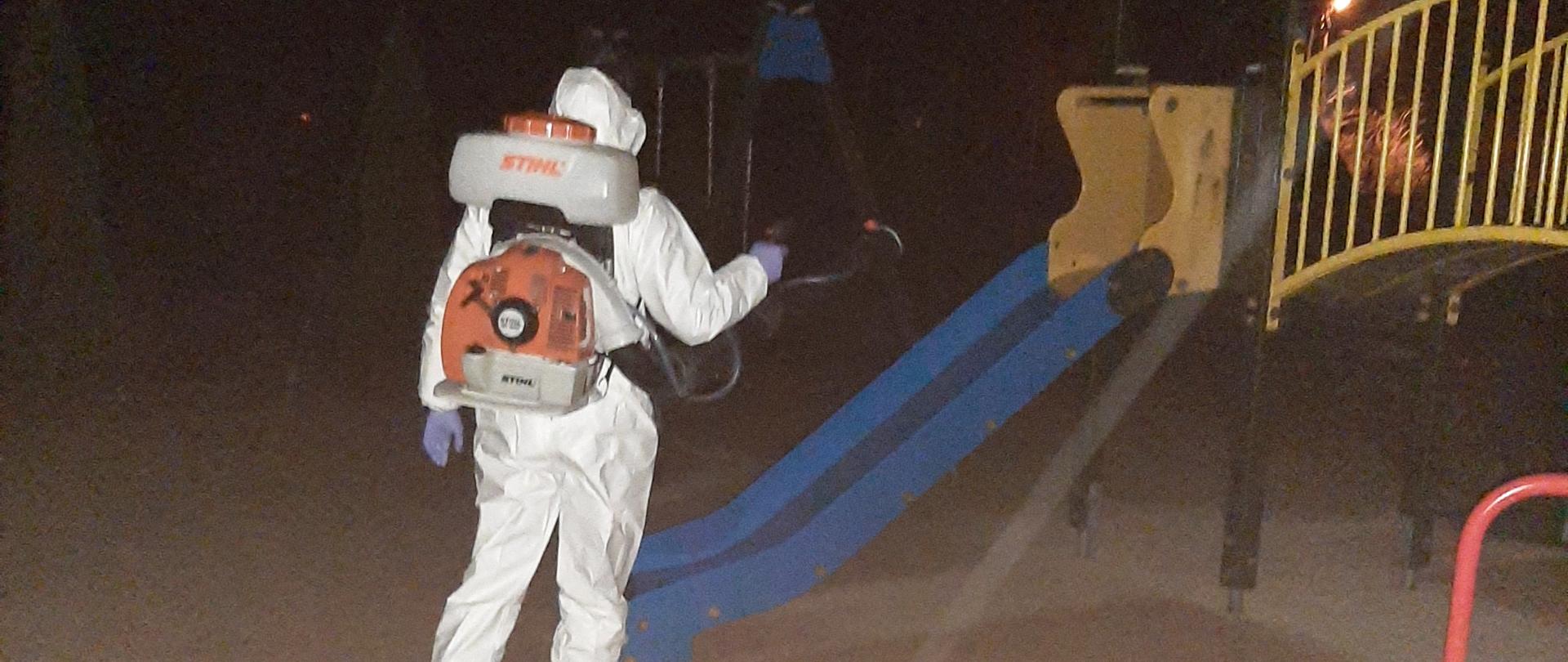 Strażak przy pomocy opryskiwacza spalinowego zabezpieczony specjalnym ubraniem dezynfekuje plac zabaw.