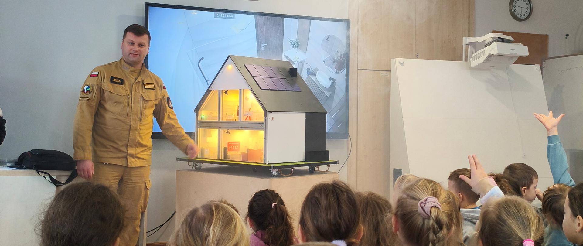 Strażak przedstawia symulator zagrożeń przed dziećmi na spotkaniu w szkolnej klasie.