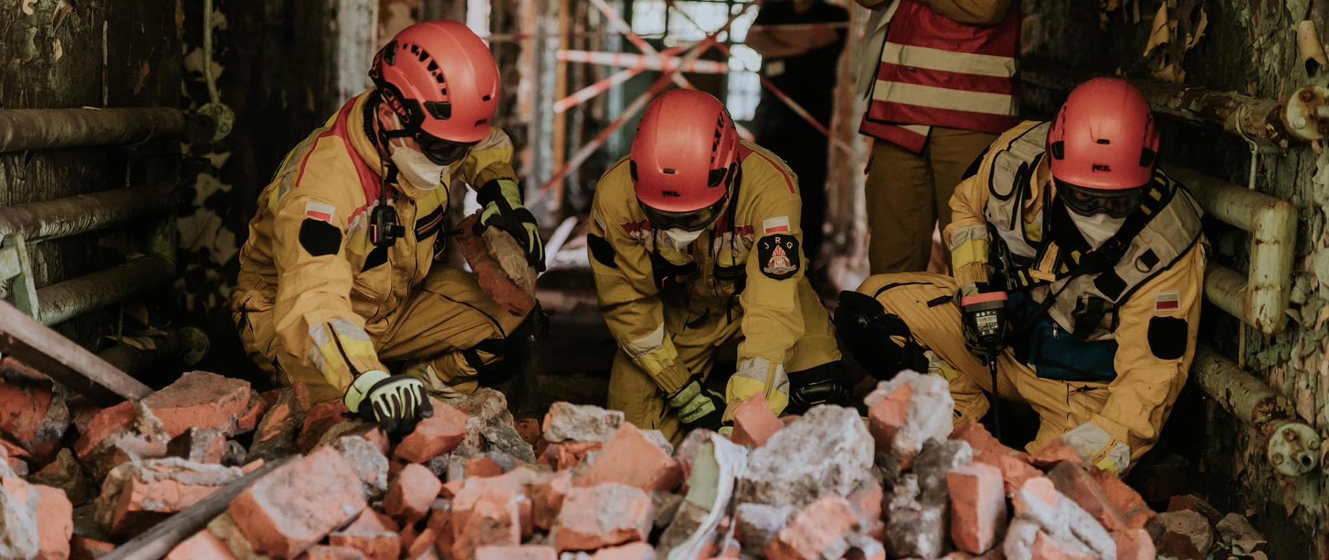 Na zdjęciu widzimy strażaków z grupy poszukiwawczo ratowniczej w piaskowych strojach prowadzących działania poszukiwawcze pod gruzami.