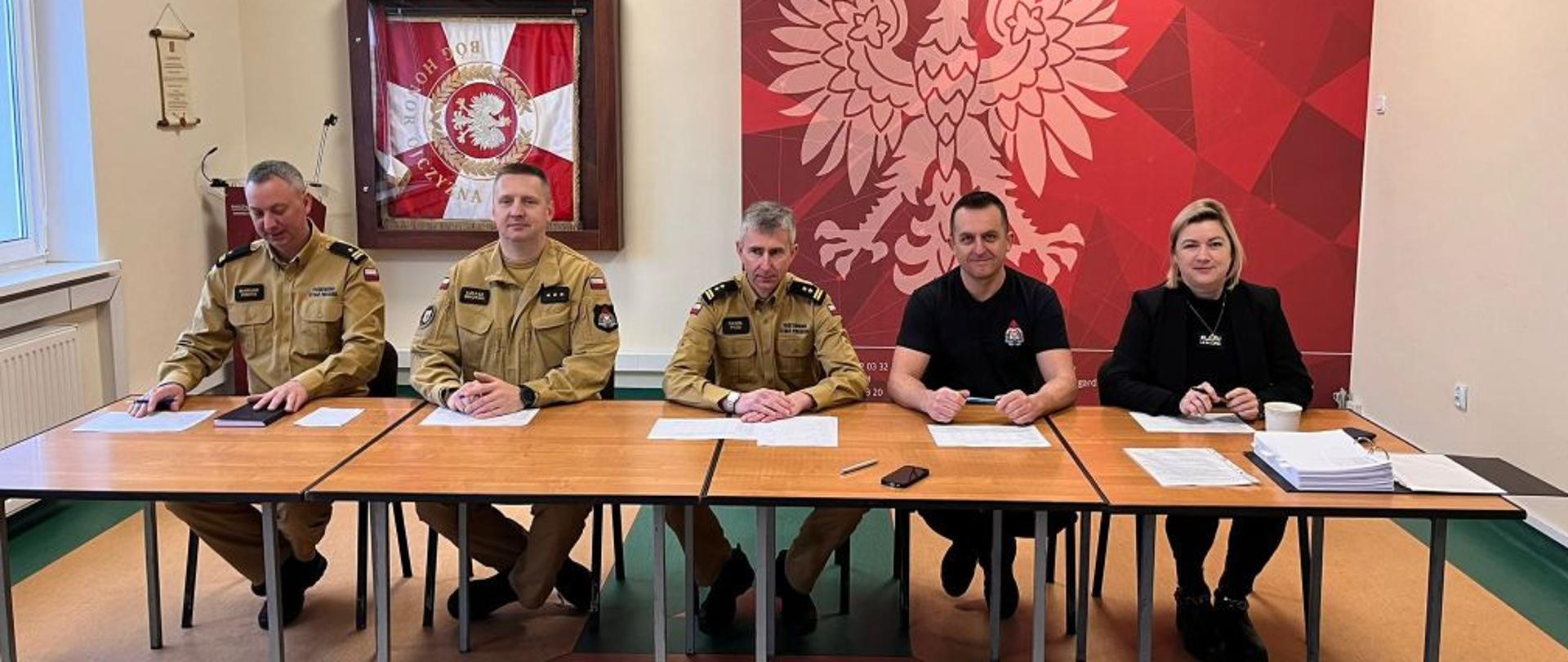 Zdjęcie wykonane na świetlicy komendy. Centralny punkt zdjęcia stanowi długi szeroki stół. Przy stole siedzi komisja rekrutacyjna, strażacy oraz kobieta, cywil. W tle ścianka reklamowa z godłem Polski.