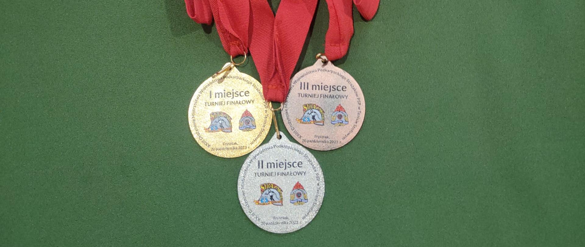 na zielony tle trzy medale z czerwonymi wstążkami za I, II, III miejsce,