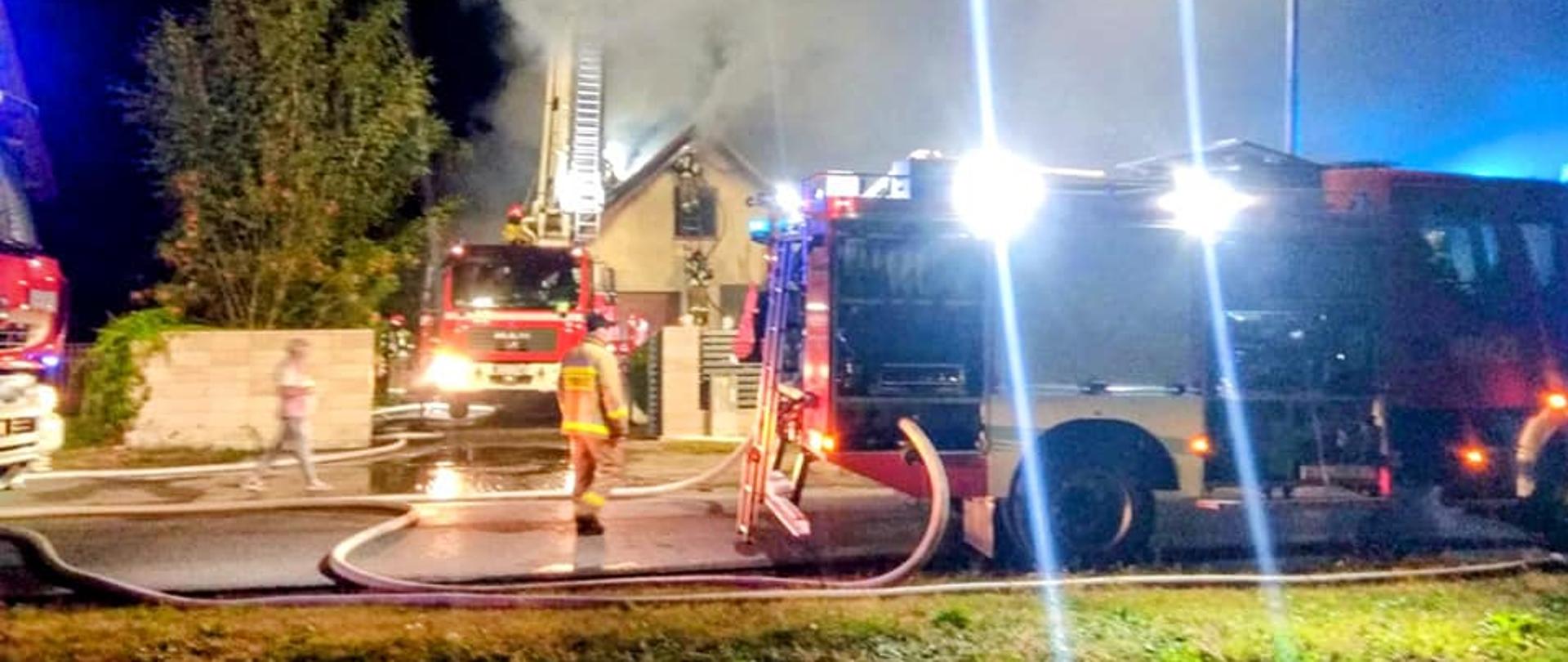 Palący się dach budynku gospodarczego. Z podnośnika przekazywane są prądy wody. Ciężki samochód oświetla teren zdarzenia. Rozwinięte węże. Wokół strażacy biorący udział w akcji.