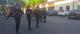 Trzech strażaków w galowych mundurach maszeruje uroczyście po placu jednostki straży pożarnej. Jeden z nich niesie złożoną biało-czerwoną flagę. W tle widoczni są inni strażacy w służbowych mundurach. Za nimi stoi czerwony wóz strażacki oraz kilka zaparkowanych samochodów osobowych. Jest słoneczny dzień.