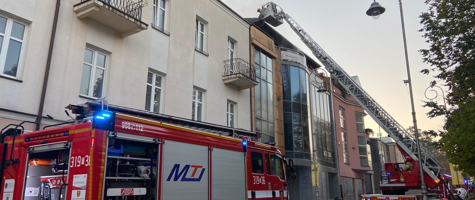 Zdjęcie przedstawia ulicę przy której doszło do pożaru kamienicy. Na jezdni stoją dwa samochody pożarnicze, w tym drabina mechaniczna rozstawiona przy kamienicy. W koszu drabiny znajdują się ratownicy. Widać zabezpieczenie i oznakowanie terenu działań. 