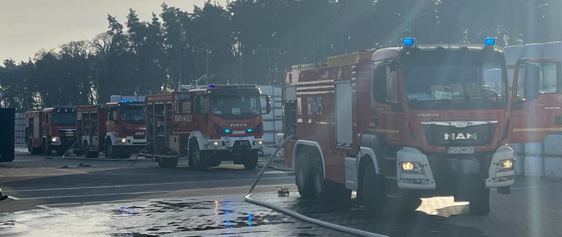 Na zdjęciu 4 czerwone samochody strażackie na włączonych sygnałach świetlnych niebieskich wraz z rozwiniętymi odcinkami pożarniczymi