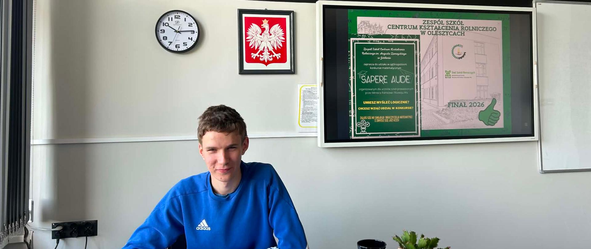 Na zdjęciu widoczny jest uczeń siedzący przy biurku podczas rozwiązywania finałowych matematycznych zadań konkursowych. Na ścianie w tle znajdują się zapisane cyfry rozwinięcia liczby π, zegar oraz godło państwowe. Fotografia przedstawia moment pracy własnej ucznia, podkreślając jego zaangażowanie i przygotowanie do konkursu.