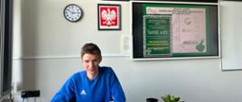 Na zdjęciu widoczny jest uczeń siedzący przy biurku podczas rozwiązywania finałowych matematycznych zadań konkursowych. Na ścianie w tle znajdują się zapisane cyfry rozwinięcia liczby π, zegar oraz godło państwowe. Fotografia przedstawia moment pracy własnej ucznia, podkreślając jego zaangażowanie i przygotowanie do konkursu.