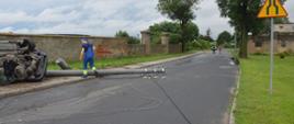 Kolizja w Trzeboszu. Auto osobowe uderzyło w słup energetyczny. Samochód leży na lewym boku. Obok na jezdni znajduje się złamany betonowy słup oraz zerwane przewody energetyczne. W tle murowane ogrodzenie gospodarstwa, a także strażak zabezpieczający drogę. Przy słupie porusza się pracownik pogotowia energetycznego.