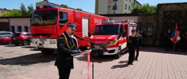 Zdjęcie wykonane w słoneczny dzień przedstawia strażaka w galowym mundurze, który stoi przy czerwonej mównicy na brukowanym placu i przemawia. Mężczyzna patrzy w dół na dokumenty leżące na mównicy, a jego prawa ręka jest uniesiona w geście salutowania lub wskazywania.
W tle, za przemawiającym strażakiem, zaparkowane są dwa czerwone wozy strażackie. Pomiędzy wozami, nieco w oddali, stoi inny strażak w galowym mundurze, patrzący prosto przed siebie. Po prawej stronie zdjęcia, w tle, widoczny jest długi, niski budynek pokryty zielonym kamuflażem, a przed nim stoi szereg strażaków w galowych mundurach oraz sztandar. W tle widoczne są również budynki mieszkalne i błękitne, bezchmurne niebo.