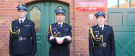 Trzech oficerów w mundurach galowych. Oficer środkowy trzyma na przedramionach złożoną flagę w kolorze biało-czerwonym. Wszyscy w postawie zasadniczej. W tle budynek z czerwonej cegły, widać drzwi i fragment wrót garażowych. Na elewacji tablica z nazwą instytucji.