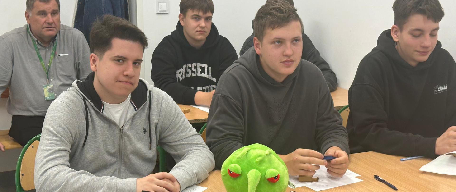 grupa uczniów siedzi w ławkach w klasie, przed nimi leży maskotka