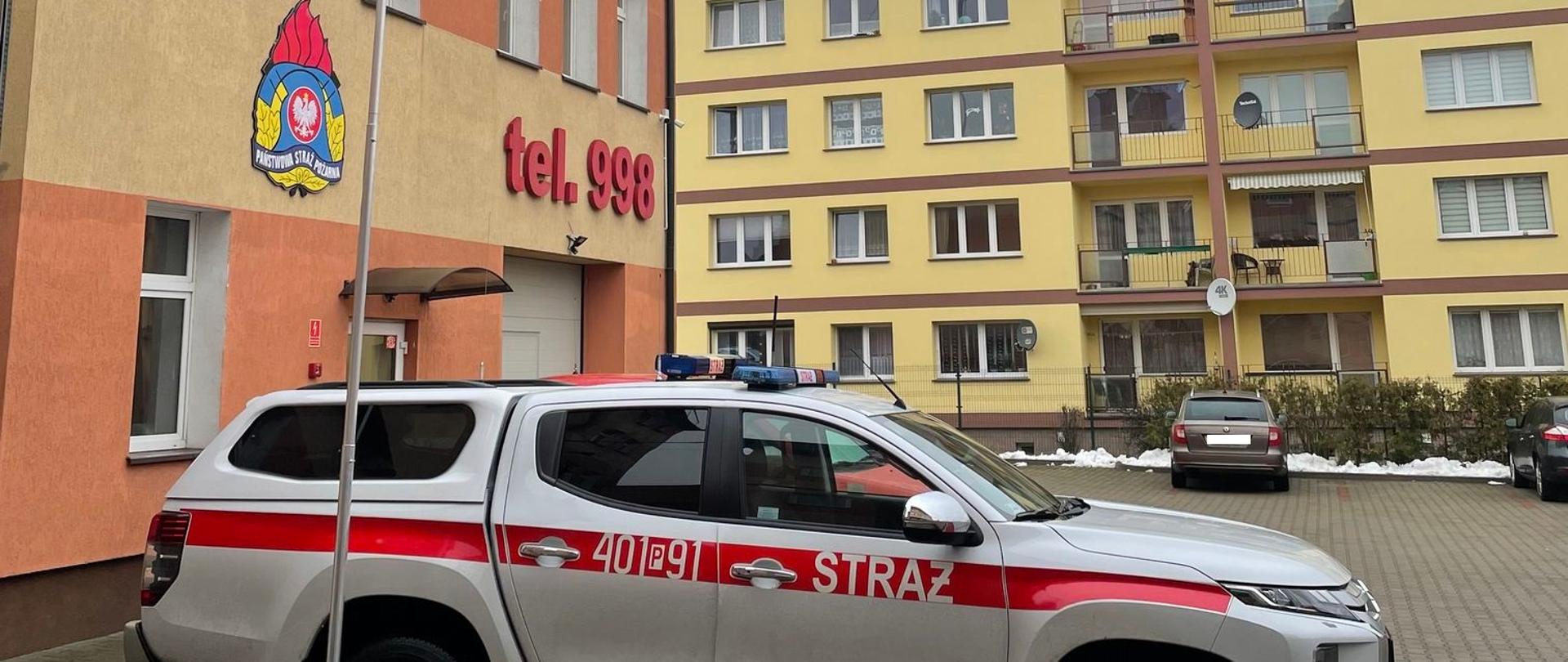 Zdjęcie przedstawia sprawiony przenośny maszt antenowy przy samochodzie strażackim.
W tle budynki.
