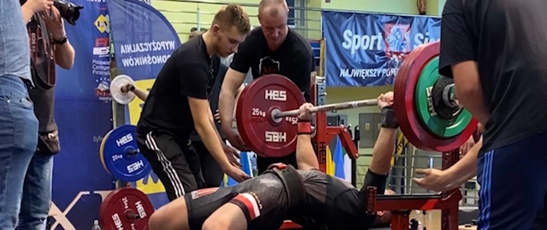 Na pierwszym planie zawodnik leżąc podnosi sztangę. Po bokach trzy osoby go asekurują.