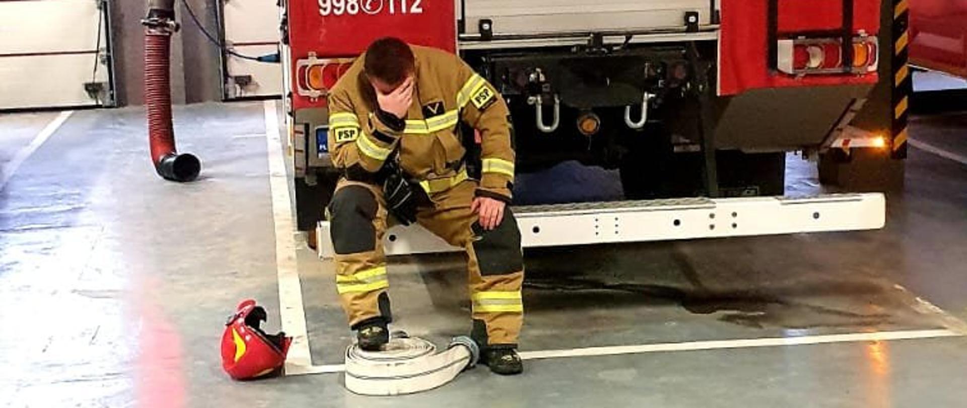 Zdjęcie przedstawia strażaka w ubraniu specjalnym koloru piaskowego. Strażak siedzi na belce czerwonego samochodu strażackiego. Zdjęcie wykonane jest w garażu Jednostki Ratowniczo Gaśniczej.