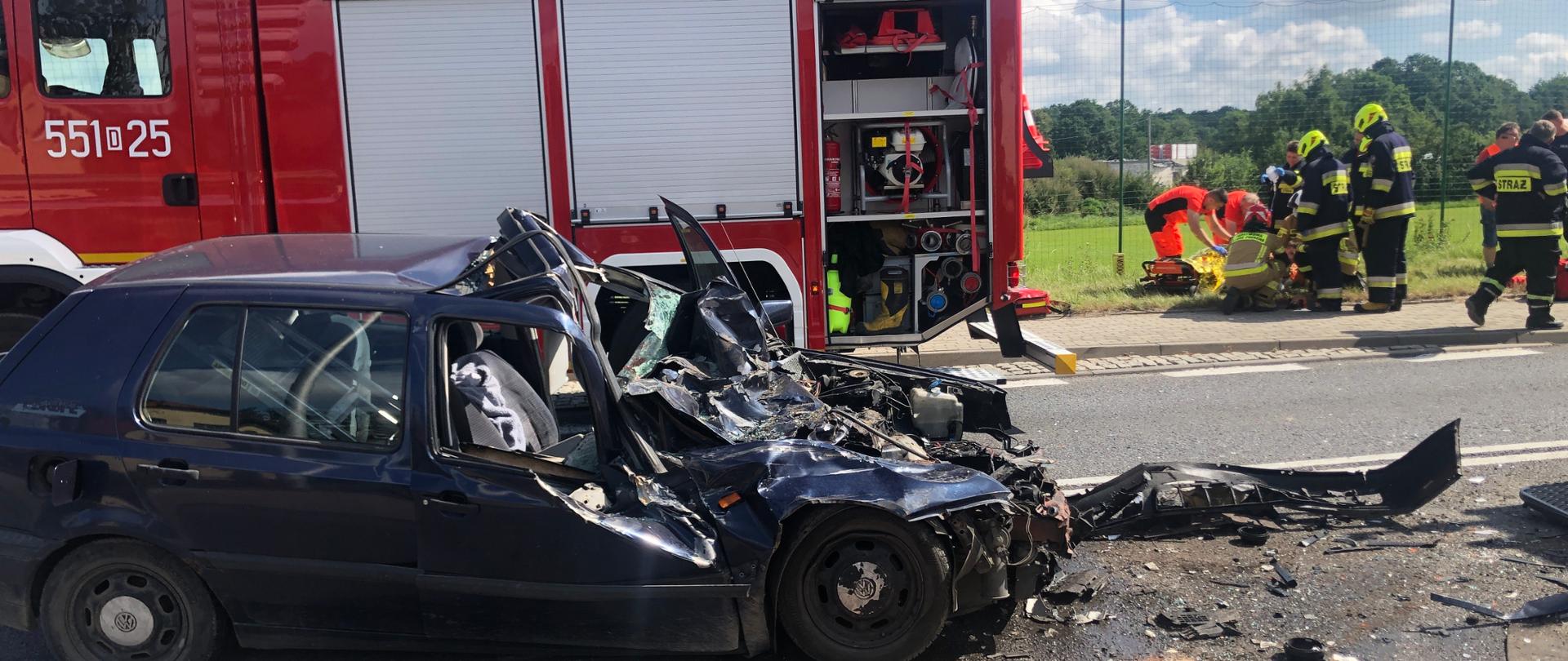na zdjęciu widać rozbity samochód osobowy, uszkodzone porozrzucane części karoserii, rozlane płyny eksploatacyjne, w tle samochód strażacki w działaniu ratowniczym oraz ratowników udzielających pierwszej pomocy osobie poszkodowanej 
