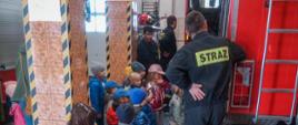 
Zdjęcie przedstawia grupę dzieci podczas zapoznania ze sprzętem strażackim w pomieszczeniu garażu na tle pojazdów strażackich koloru czerwonego.
